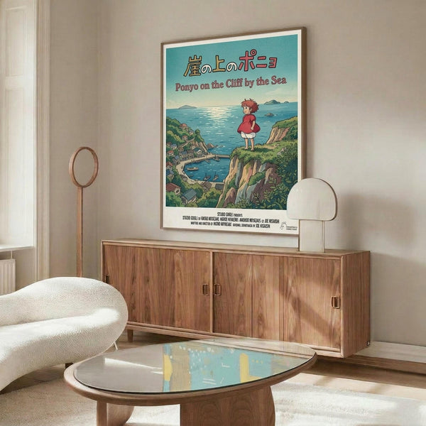 May include: Framed Ponyo movie poster hanging above wooden sideboard in modern living room. Poster shows red-dressed girl character overlooking seaside village from green cliff, with Japanese and English text on turquoise background. Room features cream curved chair, glass coffee table, and brass floor lamp against neutral walls.