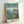 Framed poster featuring Studio Ghibli's Ponyo on the Cliff by the Sea artwork in a light wood frame. The illustration shows young Ponyo in a red dress standing on a grassy cliff overlooking a scenic coastal village with blue ocean, small boats, and houses nestled among green hills. Japanese and English text at top, Studio Ghibli credits at bottom. Frame leans against white wall casting subtle shadow.
