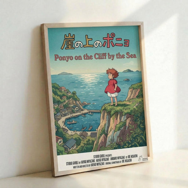 Framed poster featuring Studio Ghibli's Ponyo on the Cliff by the Sea artwork in a light wood frame. The illustration shows young Ponyo in a red dress standing on a grassy cliff overlooking a scenic coastal village with blue ocean, small boats, and houses nestled among green hills. Japanese and English text at top, Studio Ghibli credits at bottom. Frame leans against white wall casting subtle shadow.