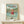 May include: Framed Studio Ghibli Ponyo poster displayed on light wood shelf against cream wall. Artwork shows red-haired child in red dress running through ocean waves with Japanese coastal town in background. Soft pastel colors in anime illustration style. Natural wood frame, small white gift box with twine bow sits in front of poster.