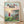 May include: Framed Studio Ghibli Ponyo movie poster print in light wood frame, leaning against white wall on wooden floor. Poster features red-haired girl in red dress overlooking coastal Japanese village with boats in harbor, mountains and sunset sky in background. Japanese and English text at top. Soft natural lighting creates gentle shadows on textured white wall.