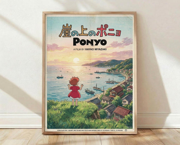May include: Framed Studio Ghibli Ponyo movie poster print in light wood frame, leaning against white wall on wooden floor. Poster features red-haired girl in red dress overlooking coastal Japanese village with boats in harbor, mountains and sunset sky in background. Japanese and English text at top. Soft natural lighting creates gentle shadows on textured white wall.