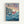 Framed poster of Studio Ghibli's Ponyo on the Cliff by the Sea featuring the red-haired fish girl character swimming in stylized blue ocean waves with white caps. A clifftop village with small houses sits on rocky cliffs in the background under a dreamy sunset sky painted in soft purples, pinks and yellows. Japanese text appears at the top with English translation below, plus film credits at the bottom. The artwork has Miyazaki's distinctive hand-drawn animation style in a light wood frame.