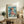 May include: Framed Studio Ghibli Ponyo poster featuring young girl with flowing brown hair in coral dress sitting by turbulent ocean waves, displayed on rustic wooden sideboard alongside fern plant, ceramic vase, and stacked books in cozy living room with beige armchair and neutral walls