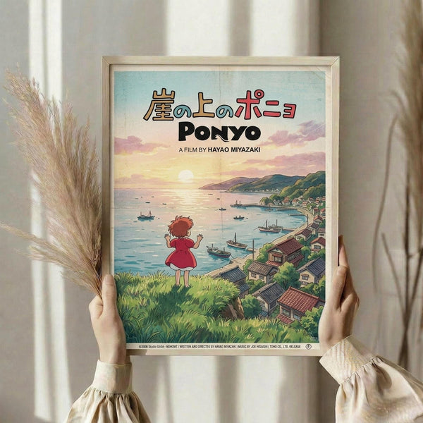 May include: Person holding framed Ponyo movie poster in light wooden frame against white curtained window, with dried pampas grass stems. Poster shows red-haired girl in red dress overlooking coastal village with boats, mountains, and sunset sky. Japanese text and "PONYO" title visible. Vintage-style anime artwork with soft pastels and detailed landscape illustration.