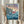 May include: Person in cream ruffled sleeves holding framed Studio Ghibli movie poster featuring red-haired character swimming in blue ocean waves near clifftop village. Japanese text at top, pastel sunset sky background. Light wooden frame, dried pampas grass visible, soft natural lighting against neutral curtained window.