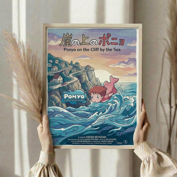 May include: Person in cream ruffled sleeves holding framed Studio Ghibli movie poster featuring red-haired character swimming in blue ocean waves near clifftop village. Japanese text at top, pastel sunset sky background. Light wooden frame, dried pampas grass visible, soft natural lighting against neutral curtained window.