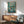 May include: Framed Ponyo movie poster hanging on white wall above wooden sideboard with lamp. Poster shows colorful underwater scene with children swimming in swirling teal water surrounded by sea creatures and bubbles. Modern living room setting with mint green upholstered chair, round wooden coffee table. Natural lighting creates geometric shadows on wall.