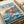 Studio Ghibli poster print featuring Ponyo, a red-haired child character in a bright red dress, joyfully running across stylized blue ocean waves. The background shows a picturesque coastal town with traditional buildings nestled on hillside cliffs. Japanese text appears at the top with English subtitle "Ponyo on the Cliff by the Sea." The artwork displays classic anime illustration style with vibrant blues, soft pastels, and detailed linework. The poster is placed on white paper with white ribb