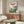May include: Framed Ponyo movie poster hanging on white wall above wooden sideboard in modern living room. Poster shows young girl in red dress overlooking coastal village with boats in harbor at sunset. Room features cream curved chair, glass-top coffee table, and warm wood furniture with natural lighting.