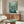 May include: Ponyo movie poster mounted on wall above wooden credenza in modern living room. Poster features animated characters surrounded by swirling turquoise water with white foam and bubbles. White upholstered chair and glass-top coffee table visible in foreground. Clean minimalist decor with neutral walls and natural lighting.