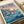 May include: Studio Ghibli-style poster print featuring red-haired Ponyo character emerging from stylized blue ocean waves. Coastal village with small houses sits on rocky cliffs under a sunset sky with pink and purple clouds. Japanese and English text at top reads "Ponyo on the Cliff by the Sea." Print displayed on light wooden surface with white rolled paper in background. Artwork shows vibrant blues, corals, and earth tones in anime illustration style.