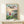 May include: Framed Studio Ghibli Ponyo movie poster displayed on light wood shelf against cream wall. Poster shows young girl in red dress overlooking coastal village with boats in harbor at sunset. Japanese and English text at top. Small white gift box sits on shelf beside the wooden-framed artwork. Soft natural lighting creates gentle shadows on minimalist setting.