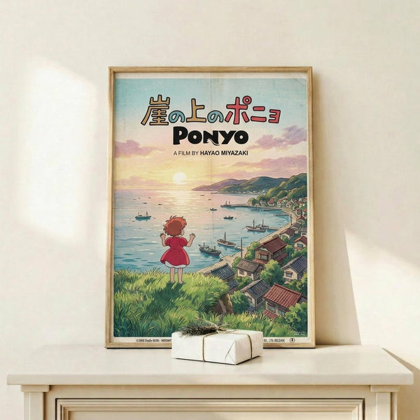 May include: Framed Studio Ghibli Ponyo movie poster displayed on light wood shelf against cream wall. Poster shows young girl in red dress overlooking coastal village with boats in harbor at sunset. Japanese and English text at top. Small white gift box sits on shelf beside the wooden-framed artwork. Soft natural lighting creates gentle shadows on minimalist setting.