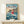 May include: Framed Studio Ghibli Ponyo movie poster displayed on light wood shelf against cream wall. Poster shows red-haired girl character jumping above ocean waves with coastal village in background, rendered in colorful anime art style. Small white wrapped gift box sits on shelf beside the wooden-framed print.