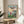May include: Framed Ponyo movie poster displayed on light wood round side table next to beige upholstered chair. Poster shows red-haired girl in red dress overlooking coastal village with boats in harbor, mountains in background under colorful sky. Japanese text at top with "PONYO" and "A FILM BY HAYAO MIYAZAKI" below. Natural lighting creates soft shadows across white walls with plant visible in corner.
