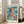 May include: Framed Studio Ghibli Ponyo movie poster leaning against white wall next to beige couch in bright living room. Poster shows young girl with flowing brown hair in pink dress sitting by turbulent ocean waves. Japanese and English text at top reads "Ponyo on the Cliff by the Sea." Artwork features vibrant blues and greens with anime-style illustration. Natural wood frame, sunlight streaming through windows.