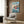 May include: Framed Studio Ghibli Ponyo movie poster mounted on wood-paneled wall above beige upholstered armchair. Poster shows red-haired girl character running through blue ocean waves with coastal village in background. Modern living room setting with brass floor lamp, geometric throw pillow, and open book on dark side table.