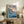 May include: Framed Studio Ghibli Ponyo movie poster displayed on rustic wooden console table beside fern plant, books, and ceramic vase. Poster shows red-haired boy swimming in blue ocean waves near clifftop village with Japanese and English text. Warm natural lighting illuminates cozy living room setting with beige armchair.