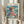 May include: Hands holding a framed Studio Ghibli Ponyo poster featuring the young red-haired character sitting by choppy ocean waves near rocky cliffs. The anime-style artwork shows Ponyo in her signature red dress against turquoise and blue seascape. Light wood frame against cream curtained background with dried pampas grass visible on left side.
