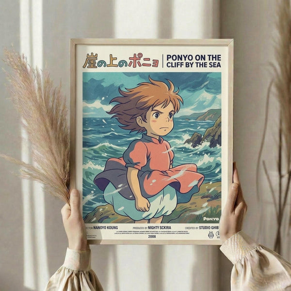 May include: Hands holding a framed Studio Ghibli Ponyo poster featuring the young red-haired character sitting by choppy ocean waves near rocky cliffs. The anime-style artwork shows Ponyo in her signature red dress against turquoise and blue seascape. Light wood frame against cream curtained background with dried pampas grass visible on left side.