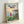 May include: Framed movie poster for Studio Ghibli's Ponyo leaning against a white wall. The poster features a young girl with red hair in a red dress standing on green grass overlooking a coastal village with boats in a harbor. The artwork shows Japanese text at the top, "PONYO" and "A FILM BY HAYAO MIYAZAKI" below, with a sunset sky in soft pastels. The poster is displayed in a light wood frame casting subtle shadows.
