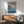 May include: Studio Ghibli-style movie poster in wooden frame hanging above mid-century modern furniture. Features animated character with red hair swimming in turquoise ocean waves near clifftop village. Japanese and English text visible. Poster displayed in minimalist room with sage green armchair, wooden credenza, white table lamp, and natural sunlight casting shadows on white walls.