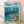 May include: A framed poster print displayed on wooden flooring against a white wall with natural shadows. The artwork features Studio Ghibli's distinctive animation style showing the character Ponyo floating in ocean water above a detailed coastal Japanese village with traditional houses, boats in the harbor, and lush green hills. The image has vibrant blues for the ocean, cloudy sky background, and includes Japanese text at the top. The poster is mounted in a natural wood frame.