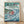 May include: Framed Studio Ghibli Ponyo movie poster in light wood frame, leaning against white wall on wooden floor. Poster features red-haired girl character swimming in stylized blue ocean waves with fish, overlooking coastal village with green hills. Japanese text at top, turquoise sky with white clouds, vibrant anime art style with Studio Ghibli film branding at bottom.