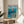 May include: Framed Studio Ghibli Ponyo poster displayed on round wooden side table next to beige upholstered chair. Artwork shows red-haired character floating in turquoise ocean water above coastal village with green hills. Japanese text and "PONYO" title visible at top. Natural wood frame, bright daylight streaming through white curtains, potted plant partially visible on left side.