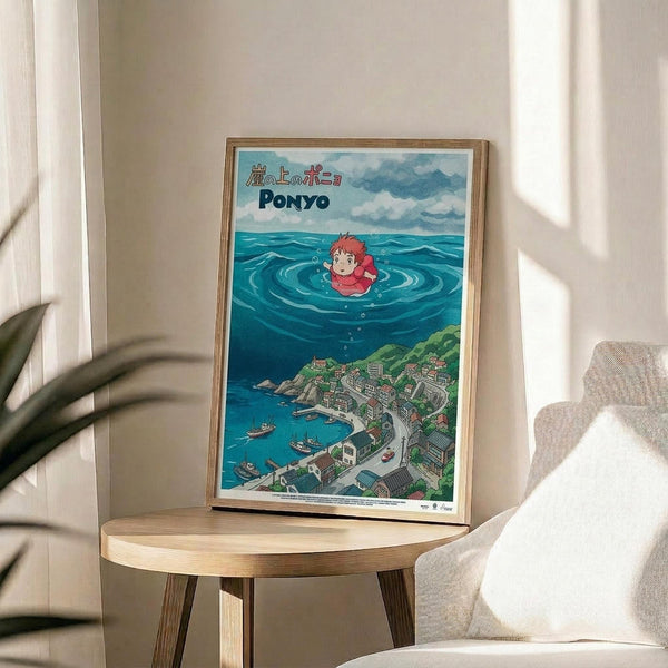 May include: Framed Studio Ghibli Ponyo poster displayed on round wooden side table next to beige upholstered chair. Artwork shows red-haired character floating in turquoise ocean water above coastal village with green hills. Japanese text and "PONYO" title visible at top. Natural wood frame, bright daylight streaming through white curtains, potted plant partially visible on left side.