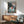 May include: Framed Ponyo movie poster displayed on white wall above wooden credenza with table lamp. Shows young girl with orange hair running through ocean waves between rocky cliffs, with house visible on clifftop. Mid-century modern room setting features green upholstered chair with wooden arms and round coffee table. Natural lighting creates shadows across minimalist interior.