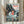 May include: Framed Studio Ghibli Ponyo poster displayed on light wooden surface with dried pampas grass beside it. Shows red-haired child character splashing in ocean waves surrounded by rocky coastal cliffs, house perched on clifftop, and sea creatures underwater. Vintage-style illustration with soft blues, greens, and coral tones in thin gold frame against neutral curtained background.