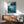 May include: Framed Ponyo movie poster displayed above mid-century modern wooden credenza in minimalist living room. Poster shows underwater scene with character swimming near rocky coastline in blue-green tones. Gray upholstered chair with wooden arms and small white table lamp visible. Natural sunlight casts geometric shadows on white wall.