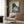 May include: Vintage-style Ponyo movie poster mounted on wood-paneled wall above beige armchair with geometric pillow. Poster shows red-haired child character splashing in ocean waves near rocky cliffs with Japanese text. Room features brass floor lamp, dark round side table with open book, and neutral modern decor.