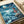 May include: Studio Ghibli-inspired poster print featuring a young girl in red dress standing on rocky cliff overlooking turbulent ocean waves under crescent moon and starry night sky. Japanese text at top reads "Ponyo on the Cliff by the Sea." Artwork shows swirling blue waters with fish below, rendered in anime illustration style. Print appears mounted or framed, photographed on light wooden surface with white fabric nearby.
