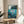 May include: Framed poster with underwater scene showing red-haired character swimming among sea plants and rocks, with cliffs above water surface. Wooden frame on round wooden side table next to beige upholstered chair, with plant shadows casting across white wall in bright natural lighting.