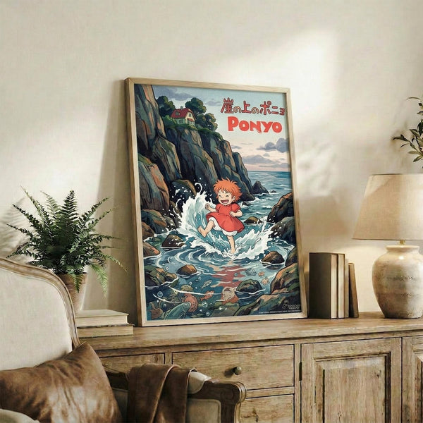 May include: A framed poster featuring the animated character Ponyo, a small red-haired child in a red dress splashing in ocean waves near rocky cliffs, with Japanese text and "PONYO" in red letters at the top. The wooden-framed artwork is displayed on a rustic wood sideboard alongside books, a ceramic vase, and a fern plant, with a cream upholstered chair visible in a cozy living room setting.
