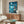 May include: Studio Ghibli-style anime poster showing a young girl in red dress standing on rocky cliff overlooking swirling blue ocean waves under starry night sky with full moon. Poster displays Japanese text and "Ponyo on the Cliff by the Sea" title. Mounted above mid-century modern wooden credenza with brass handles in minimalist living room setting with white upholstered chair, oval glass coffee table, and cream walls.
