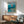 May include: Framed Studio Ghibli Ponyo poster displayed on white wall above wooden sideboard with lamp. Shows red-haired Ponyo character swimming in ocean waves above coastal Japanese town. Mid-century modern living room setting with teal armchair, round coffee table, and natural lighting creating geometric shadows on wall.