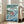 May include: Framed Studio Ghibli Ponyo movie poster in light wood frame, leaning against white wall next to beige sofa in bright living room. Poster shows red-haired girl character riding large ocean wave with fish, green hills and village in background, Japanese text at top. Turquoise and blue water colors dominate the anime-style artwork.