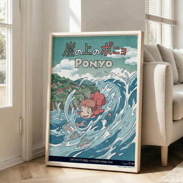 May include: Framed Studio Ghibli Ponyo movie poster in light wood frame, leaning against white wall next to beige sofa in bright living room. Poster shows red-haired girl character riding large ocean wave with fish, green hills and village in background, Japanese text at top. Turquoise and blue water colors dominate the anime-style artwork.