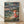 May include: Framed Studio Ghibli Ponyo movie poster print leaning against white textured wall on light wood floor. Artwork shows red-haired child in red dress sitting on rocky coastline holding glass jar, with stormy ocean waves, dramatic cloudy sky, and hillside village in background. Natural wood frame, vintage illustration style with muted greens, blues, and warm earth tones.