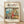 May include: Framed Studio Ghibli poster showing Ponyo character in red dress cooking in cozy kitchen with ocean cliff view through window. Light wood frame against white textured wall on wooden floor with natural lighting and shadows. Vintage anime art style with warm colors and detailed interior scene.