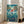 May include: Framed Studio Ghibli Ponyo movie poster in light wood frame leaning against white wall next to beige sectional sofa. Poster features red-haired Ponyo character surfing on ocean waves with coastal village, lighthouse, and boats in background. Underwater scene shows colorful fish and sea creatures. Bright blues, teals, and coral colors with Japanese text and English title at bottom.