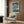 May include: Framed Studio Ghibli Ponyo movie poster mounted on wood-paneled wall above cream upholstered armchair with geometric pillow. Poster shows animated characters riding turbulent ocean waves during storm with dark cloudy sky and coastal buildings. Modern reading corner with brass floor lamp and small round table with open book. Natural lighting from window with sheer curtains.