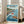 May include: Framed poster in light wood frame leaning against white wall next to beige sofa in bright room with windows. Artwork shows anime-style scene with red-haired girl emerging from ocean wave next to small wooden boat with boy inside, coastal landscape with lighthouse and houses in background, blue sky with white clouds. Japanese and English text at top and bottom of colorful illustration.