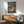 May include: Framed Studio Ghibli Ponyo movie poster mounted on white wall above wooden sideboard with white table lamp. Poster shows red-haired child character sitting on rocky coastline with ocean waves and cliffside village in background. Mid-century modern living room setting with sage green upholstered armchair, round coffee table, and natural sunlight creating geometric shadows on wall.