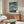 May include: Framed Studio Ghibli Ponyo movie poster displayed above a wooden mid-century modern credenza in a minimalist living room. The colorful anime artwork shows characters in stormy ocean waves with Japanese text. White curved chair and glass coffee table visible in foreground. Clean aesthetic with beige walls and natural lighting.