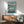 May include: Framed Studio Ghibli Ponyo movie poster displayed above a mid-century modern wooden sideboard in a minimalist living room. The colorful artwork shows red-haired Ponyo emerging from ocean waves near a small fishing boat, with coastal cliffs and lighthouse in background. Natural sunlight streams across white walls. Teal upholstered chair with wooden arms and small white table lamp visible in corner.